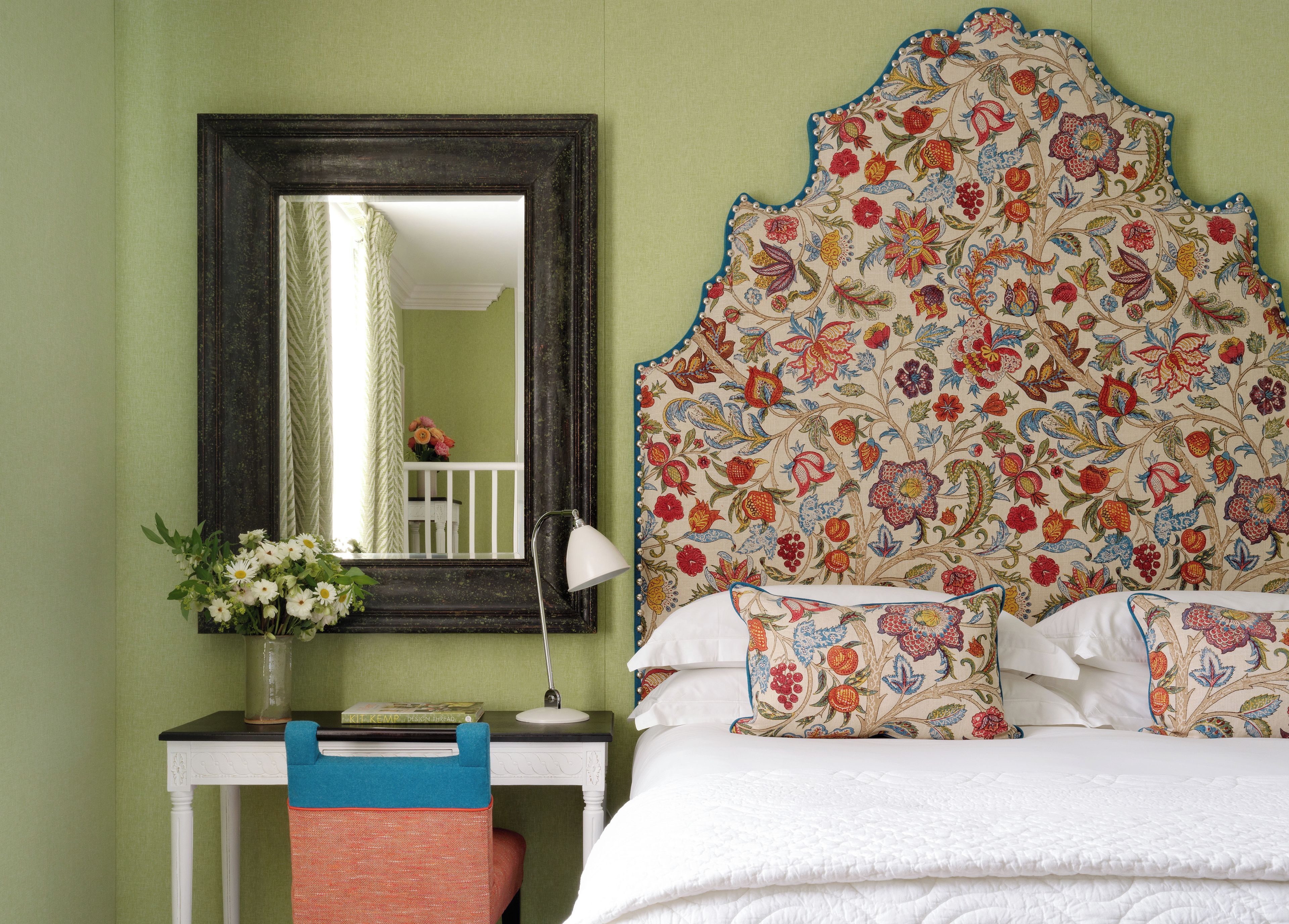 A bedroom with a bed, a bedside table, a lamp, a mirror, and a floral-patterned headboard.