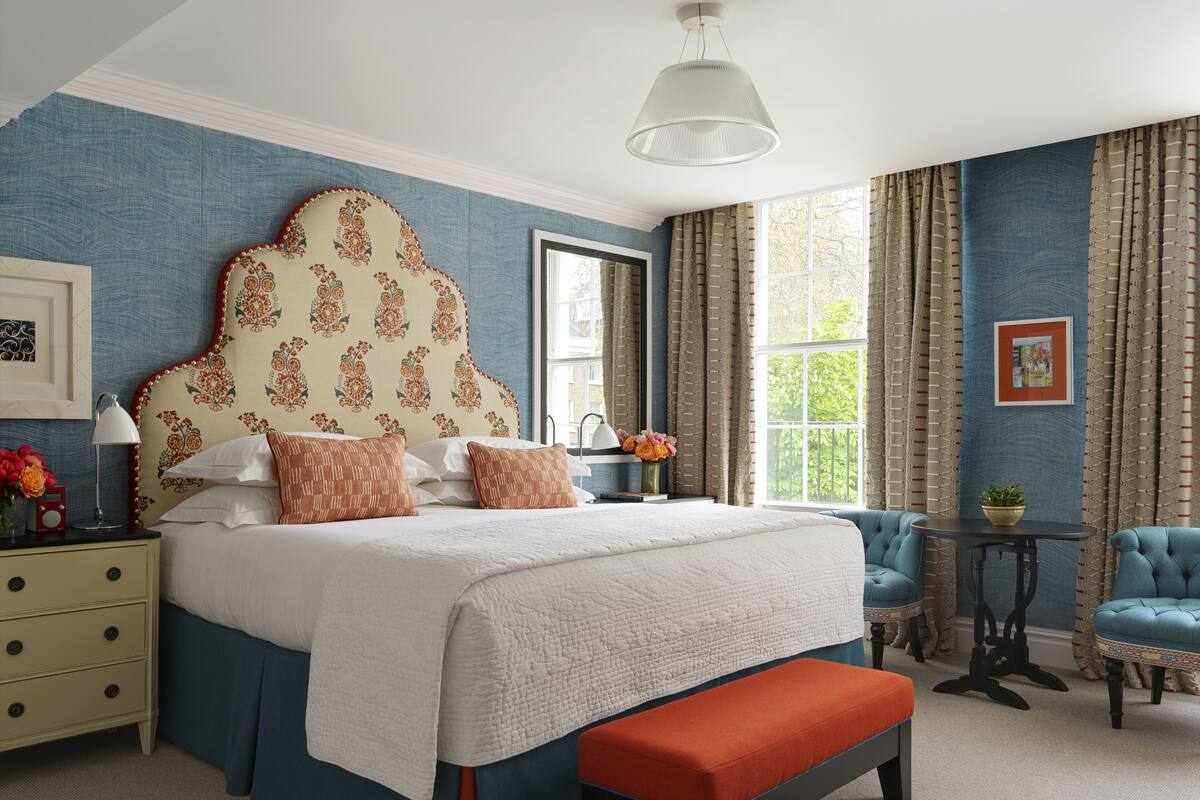 A bedroom with a large bed, a bedside table with a lamp, a bench, and a small table with a plant.