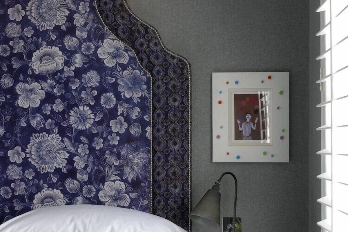 A bedroom with a large floral-patterned headboard, a white bedspread, and a matching pillow. There is a bedside table with a lamp and a small plant. A framed picture hangs on the wall.