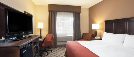 A hotel room with a bed, a desk with a television, a chair, a lamp, and a window with curtains.