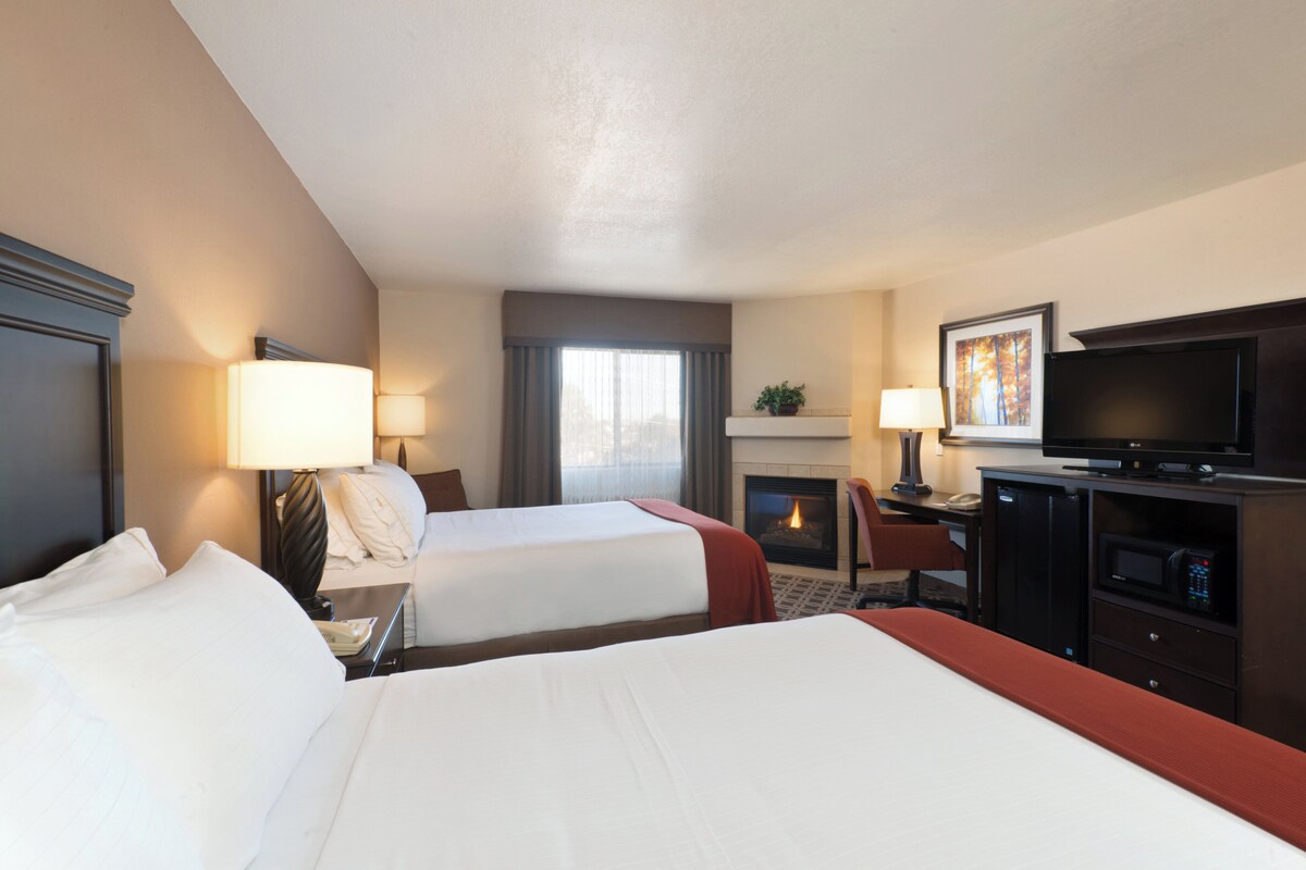 A hotel room with two beds, a TV, a fireplace, and a desk with a lamp.