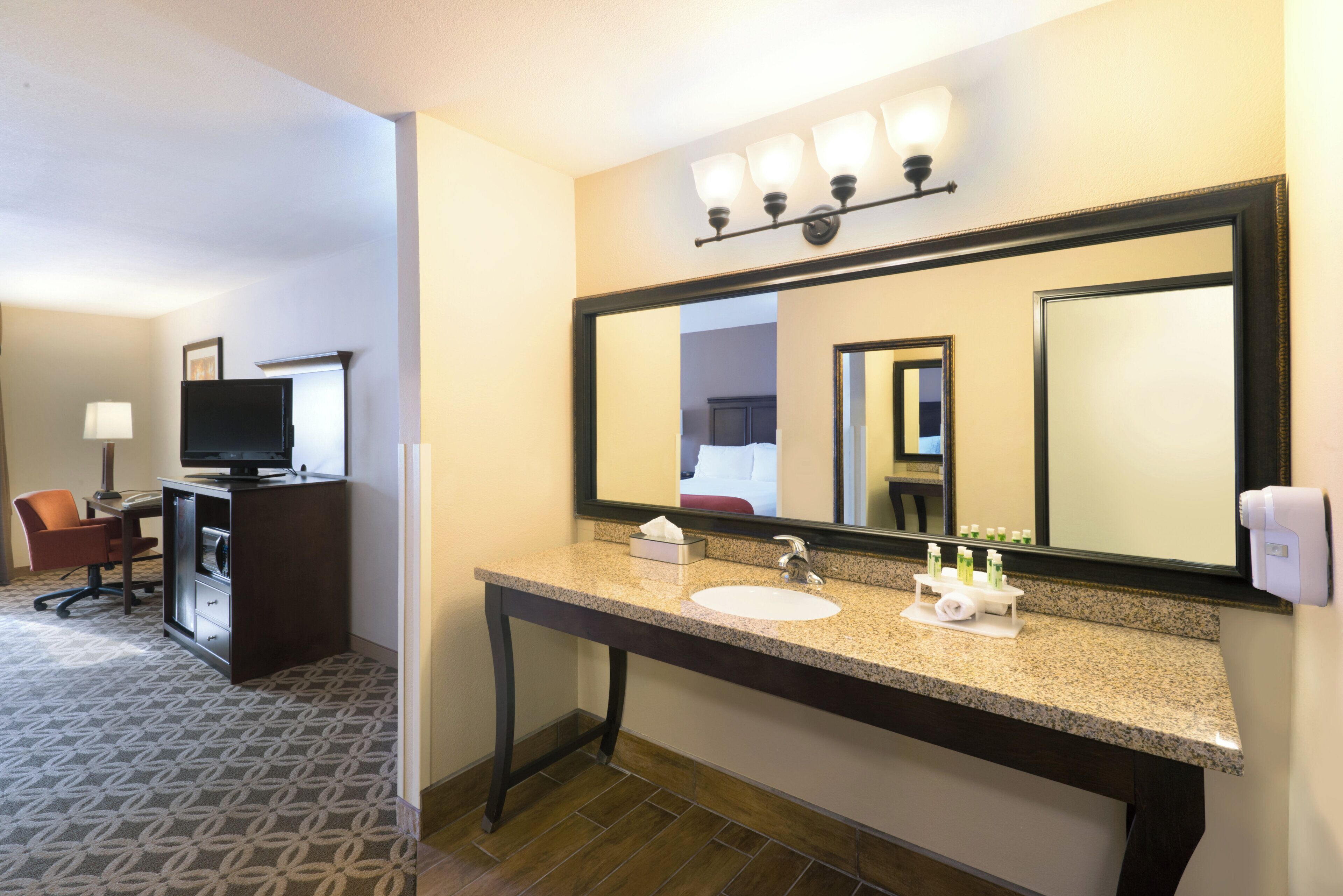 A hotel bathroom with a large mirror, a sink, a vanity, and a television in the adjacent room.