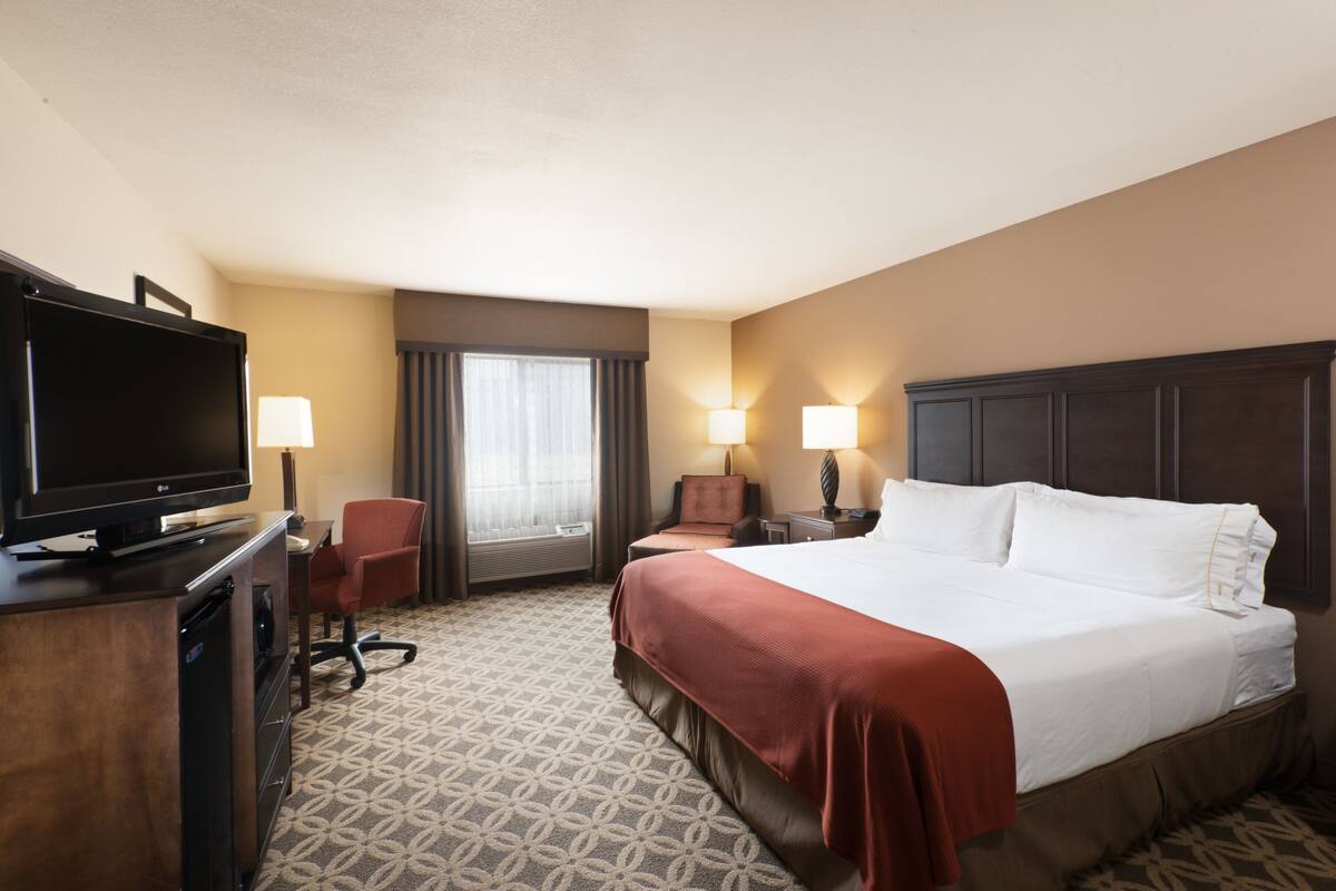 A hotel room with a large bed, a desk with a television, two bedside lamps, a chair, and a window with curtains.