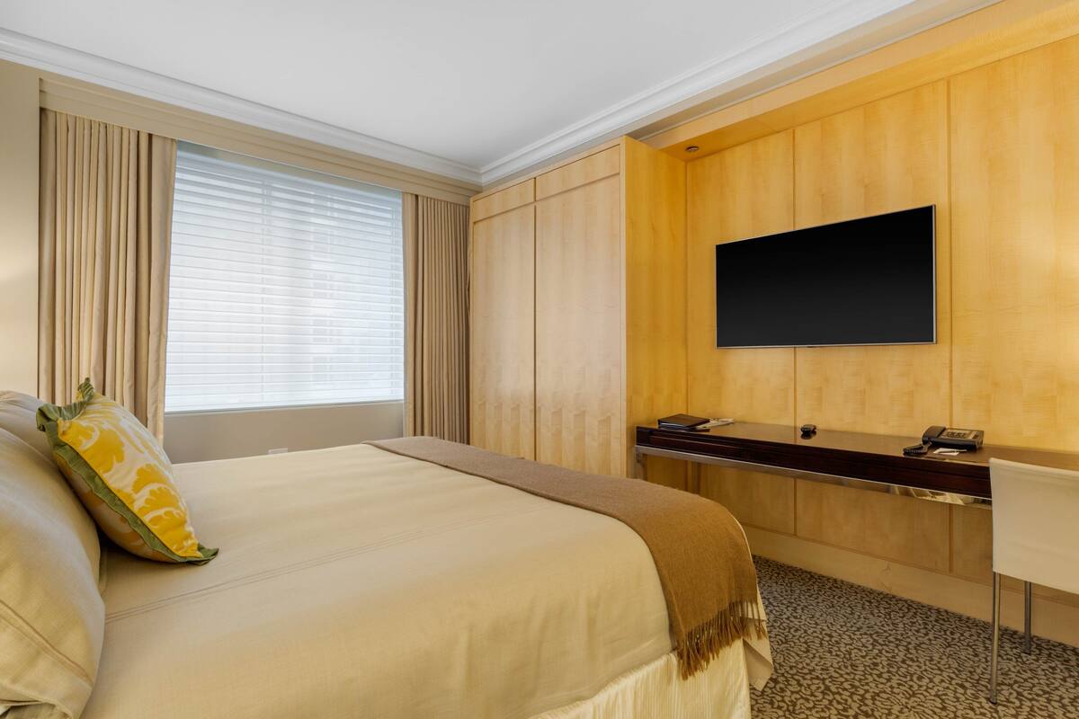 Quarto de hotel com uma cama grande, uma escrivaninha e uma televisão de tela plana, além de uma janela com cortinas.