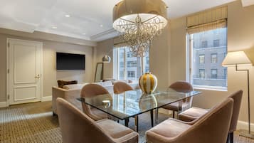 A modern dining room with a glass table, four chairs, a chandelier, a large vase, and a television on the wall.