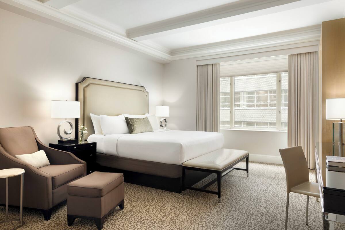 Quarto de hotel com cama grande, uma cadeira, escrivaninha e janela com cortinas.
