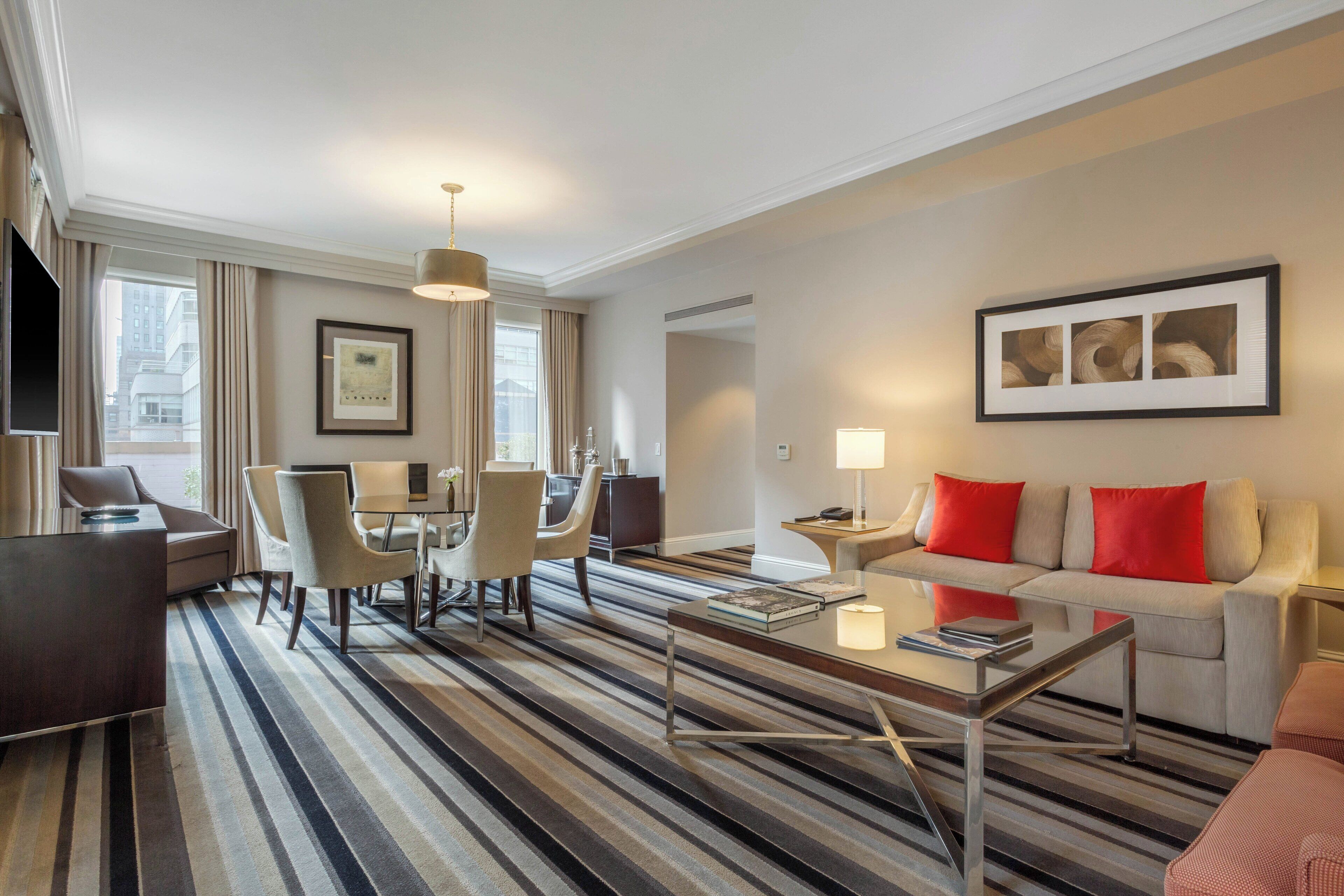 A hotel room with a striped carpet, a sofa with red cushions, a dining area with chairs and a table, a TV, and artwork on the walls.
