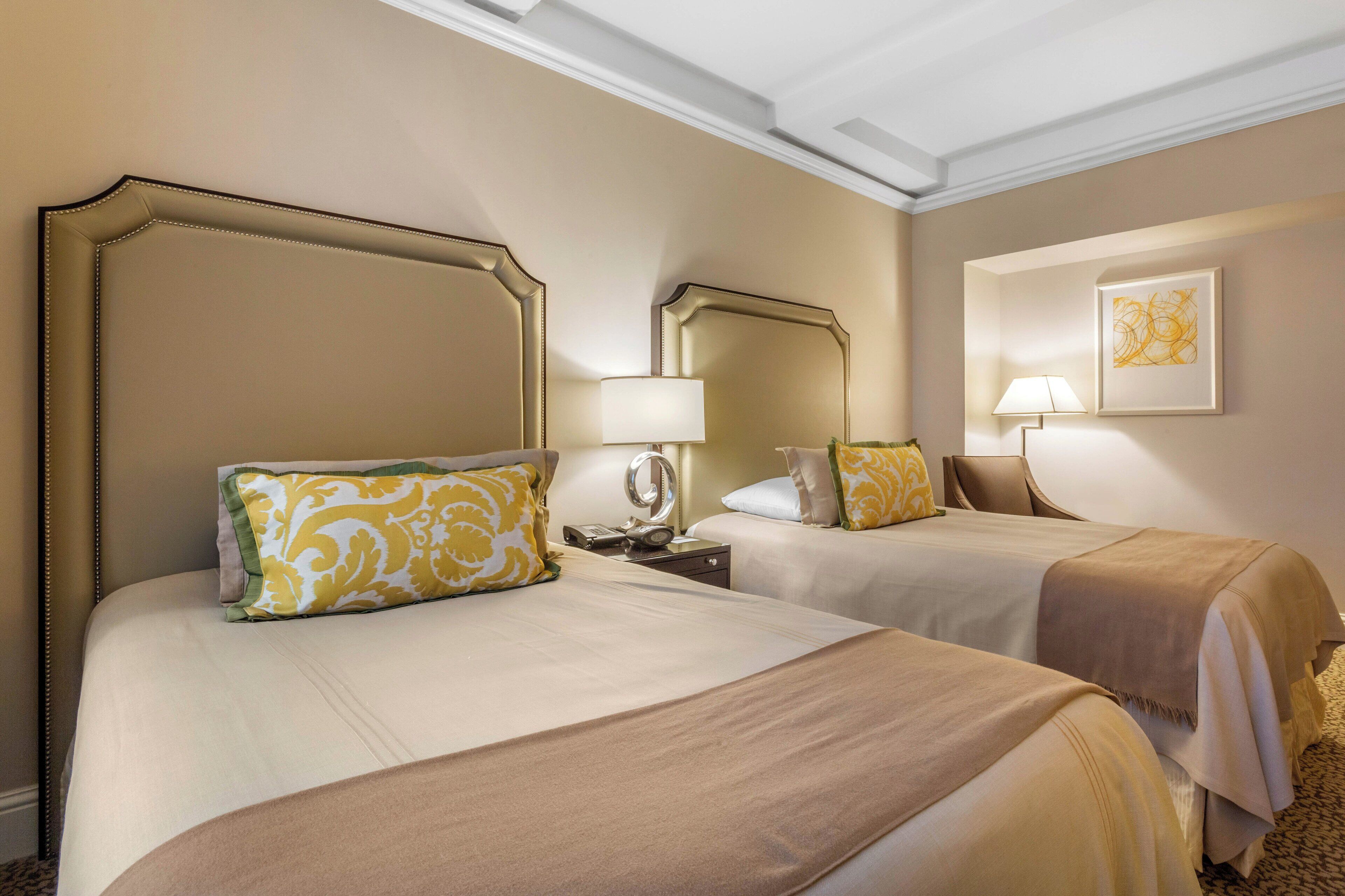 A hotel room with two beds, each with a headboard, a bedside table with a lamp, and a framed artwork on the wall.