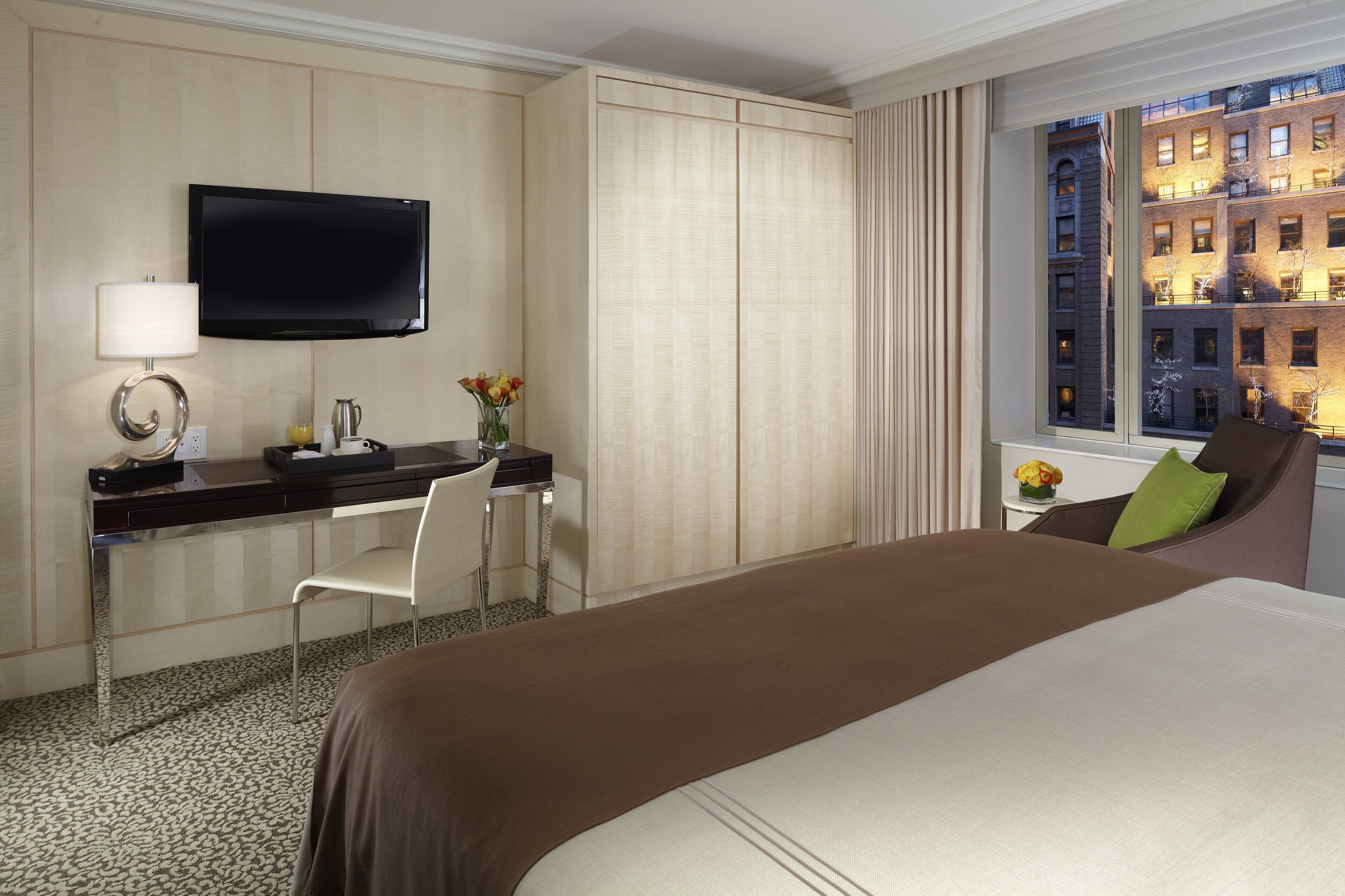 A hotel room with a bed, a desk with a chair, a television, and a window with a city view.