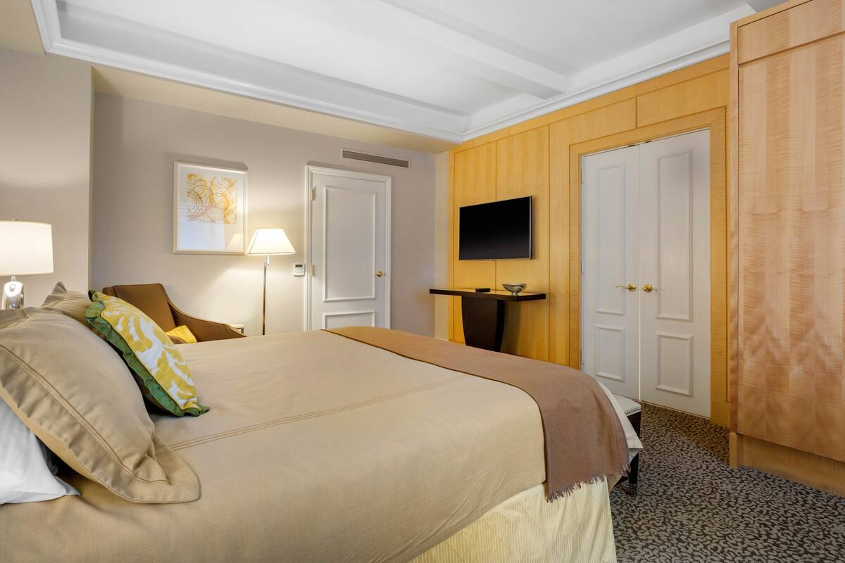 Quarto de hotel com uma cama grande, uma televisão na parede, um guarda-roupa de madeira e uma mesa de cabeceira com uma luminária.