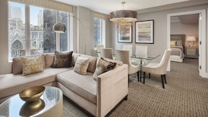 A modern hotel room with a large window offering a city view, a cozy seating area with a sofa and chairs, a glass dining table, and a chandelier.