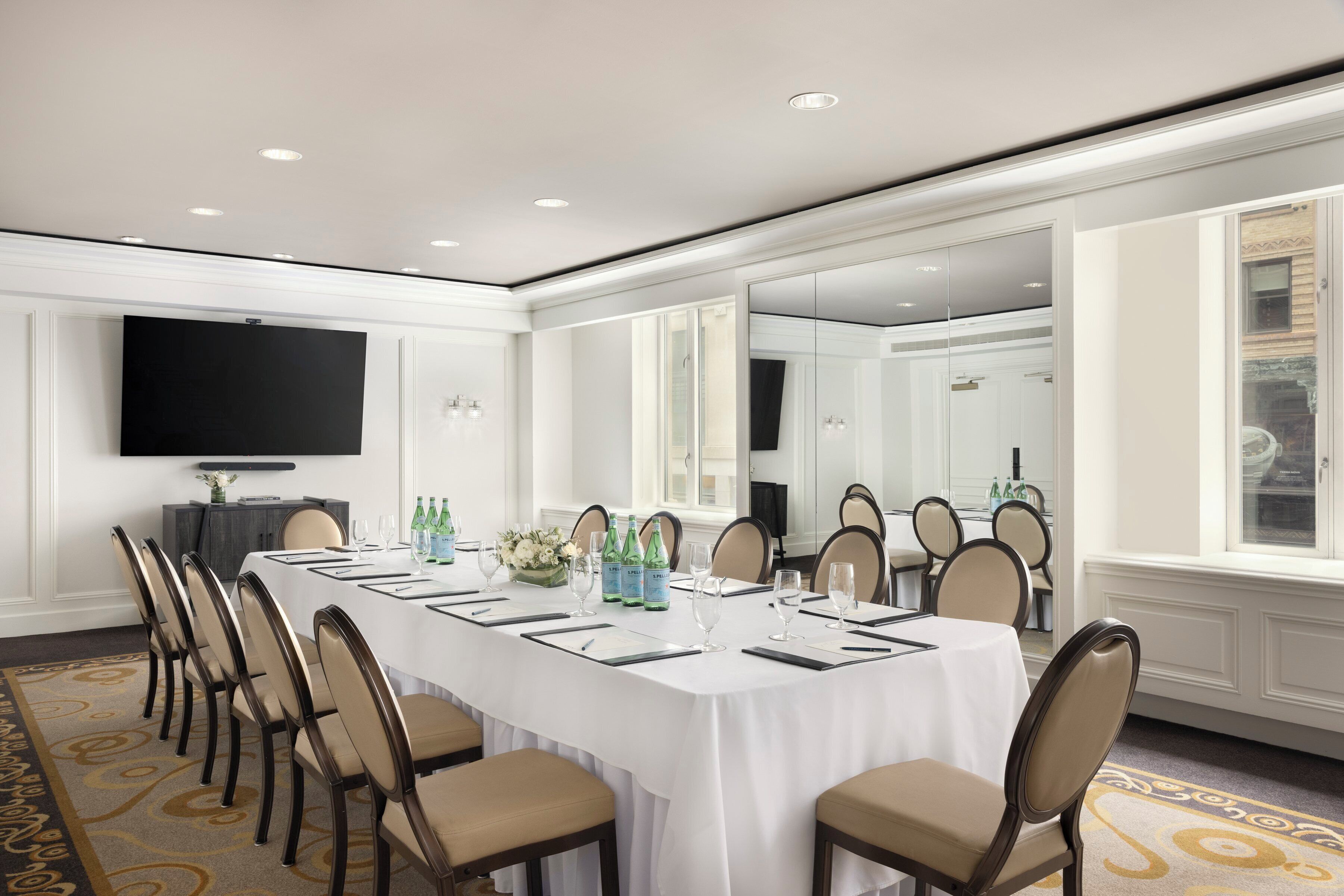 A conference room with a long table, chairs, a flat-screen TV, and a large mirror.