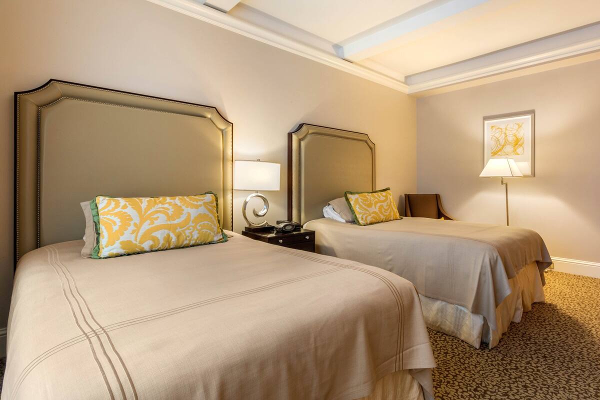 Quarto de hotel com duas camas, cada uma com cabeceira, mesa de cabeceira, abajur e um quadro na parede.