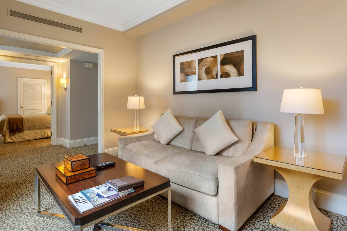 Quarto de hotel com um sofá bege, uma mesa de centro de madeira com revistas, um quadro na parede e duas luminárias de cabeceira.
