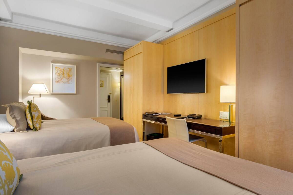 Quarto de hotel com duas camas, uma escrivaninha, uma cadeira, televisão de tela plana e uma luminária.