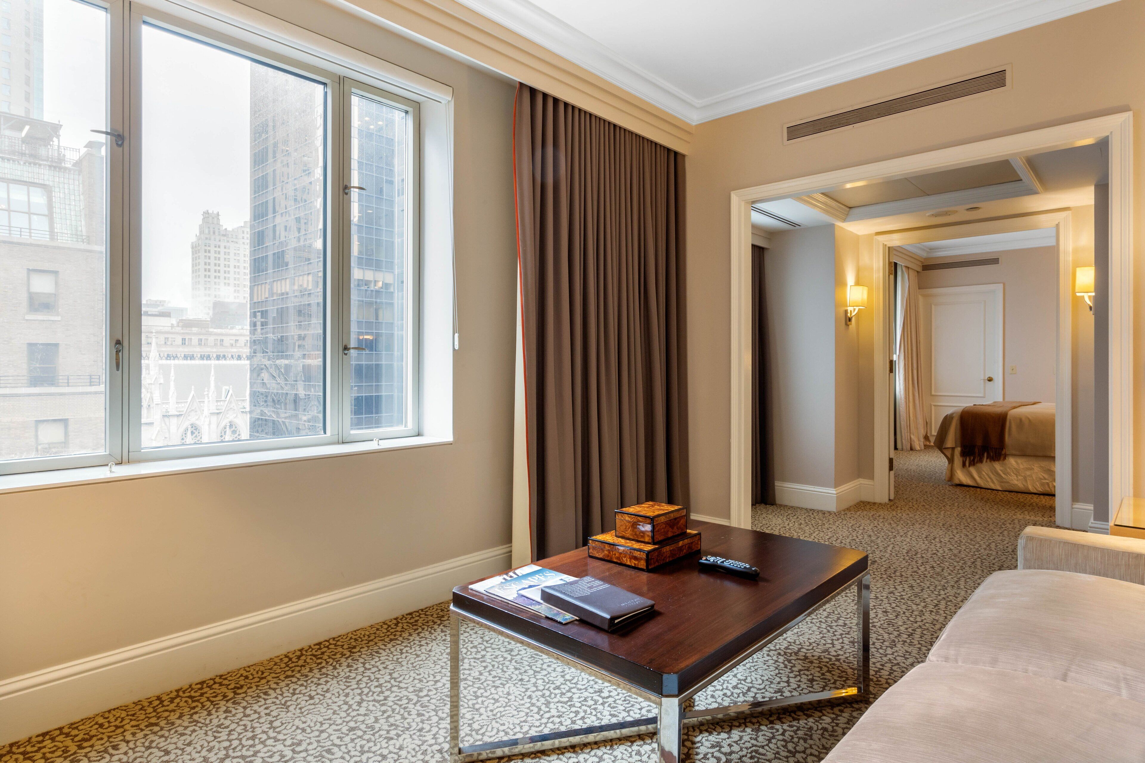 A hotel room with a large window, a wooden coffee table with a remote control, a small wooden box, and a bed in the background.