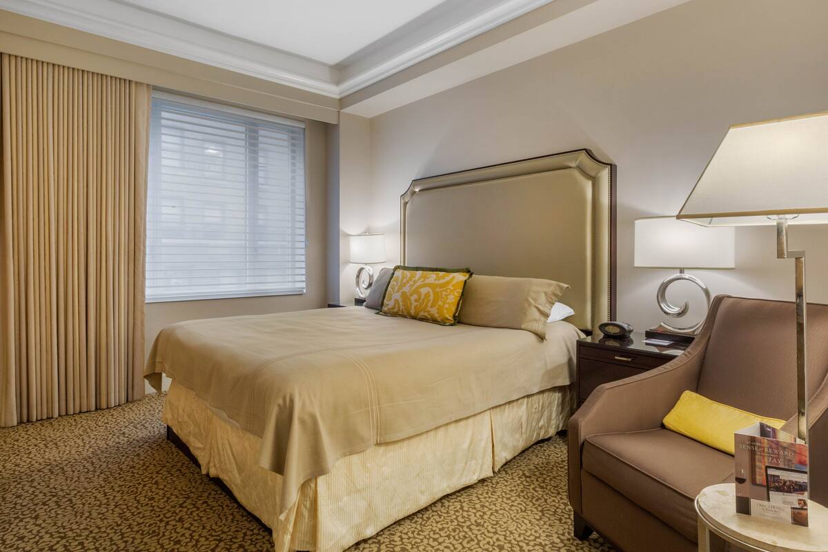 Quarto de hotel com uma cama grande, mesa de cabeceira, uma cadeira, abajur e janela com persianas.