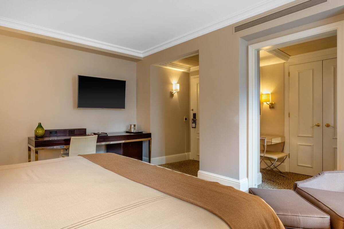 Quarto de hotel com uma cama grande, uma escrivaninha com cadeira, televisão de tela plana na parede e uma porta que dá para outro quarto.
