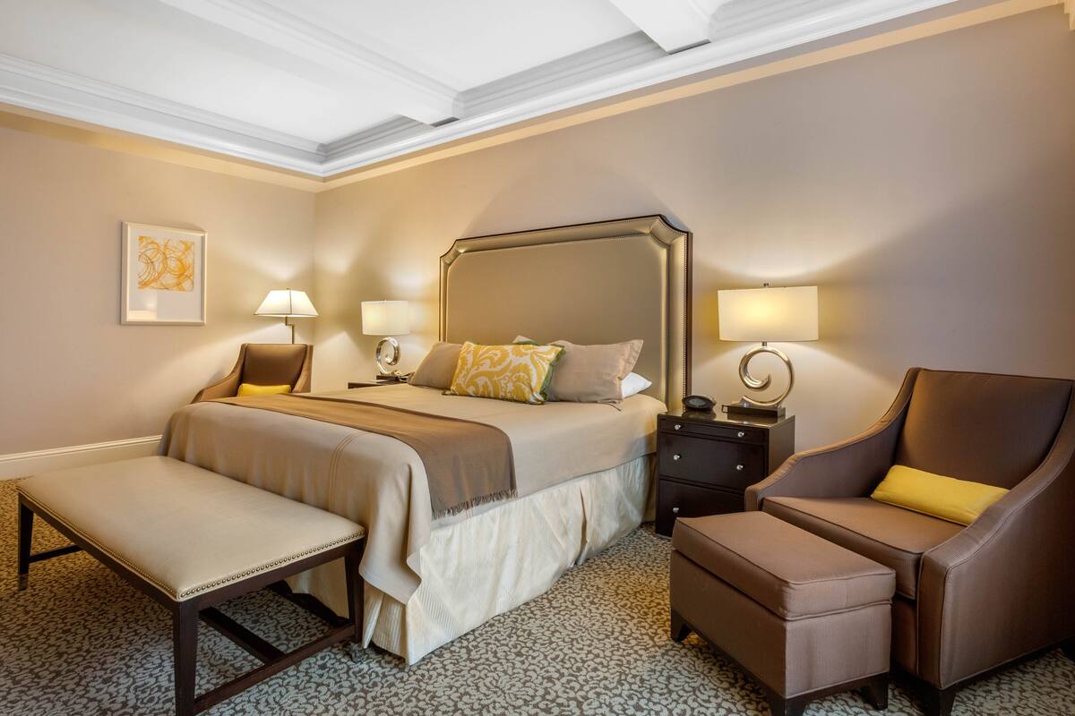 Quarto de hotel com uma cama grande, duas mesas de cabeceira com luminárias, uma cadeira, um banquinho e um tapete estampado.