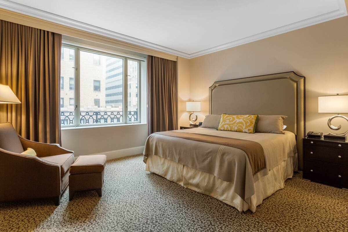 Quarto de hotel com uma cama grande, uma cadeira, cômoda e janela com cortinas.