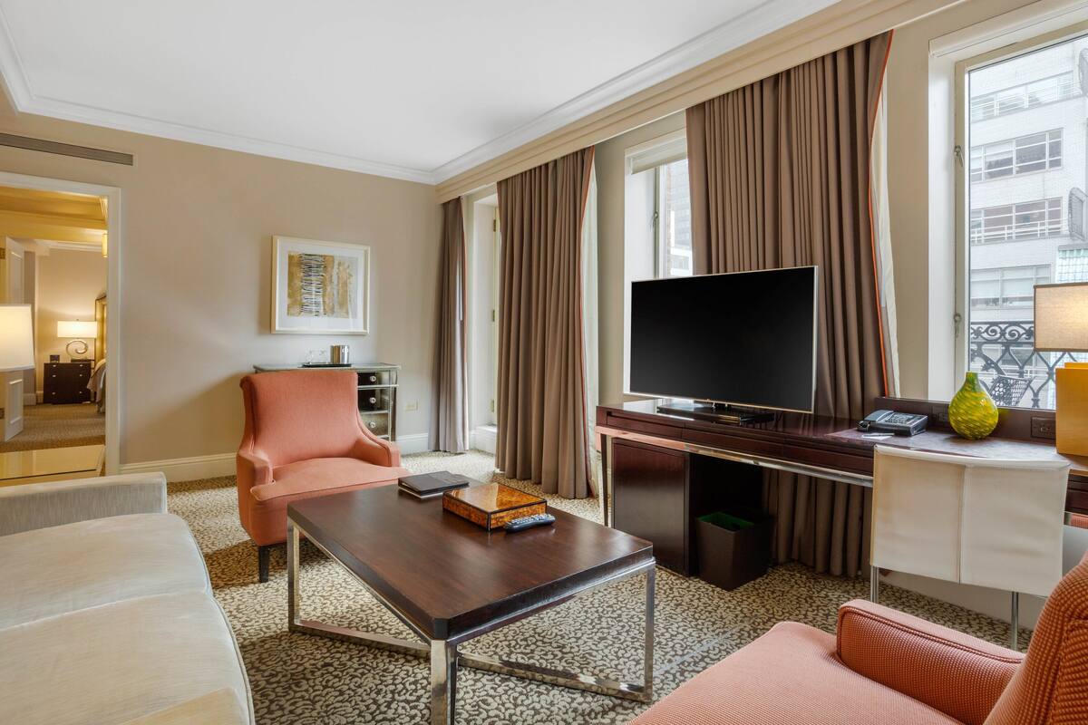 Quarto de hotel com televisão de tela plana, escrivaninha, cadeira, sofá e janela com cortinas.