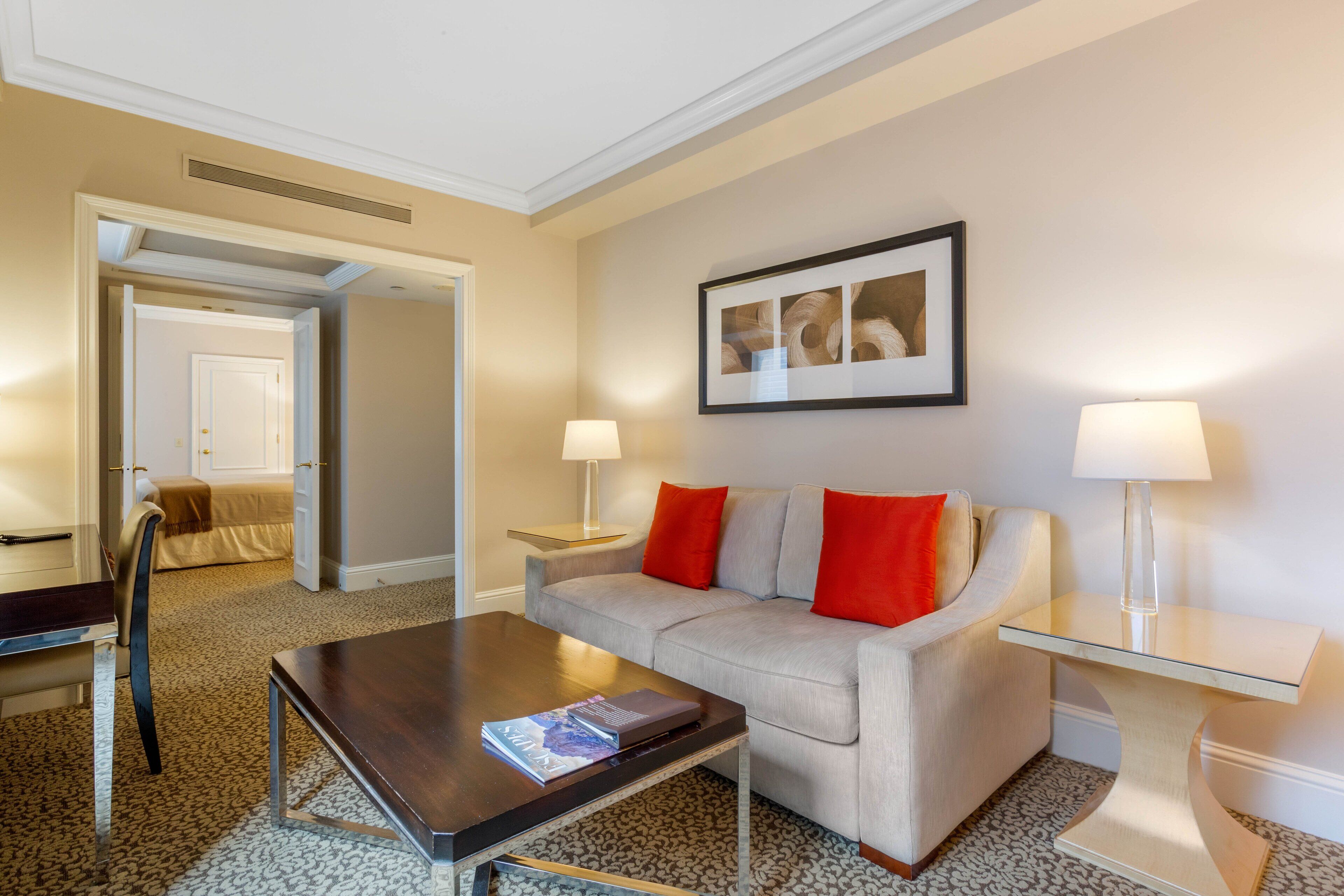 Een hotelkamer met een bank, een salontafel, een nachtkastje met een lamp, een lijst met een foto aan de muur, en een deur naar een andere kamer.