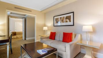 Habitación de hotel con sofá, mesa de centro, mesita de noche con lámpara, un cuadro en la pared y una puerta que da a otra habitación.