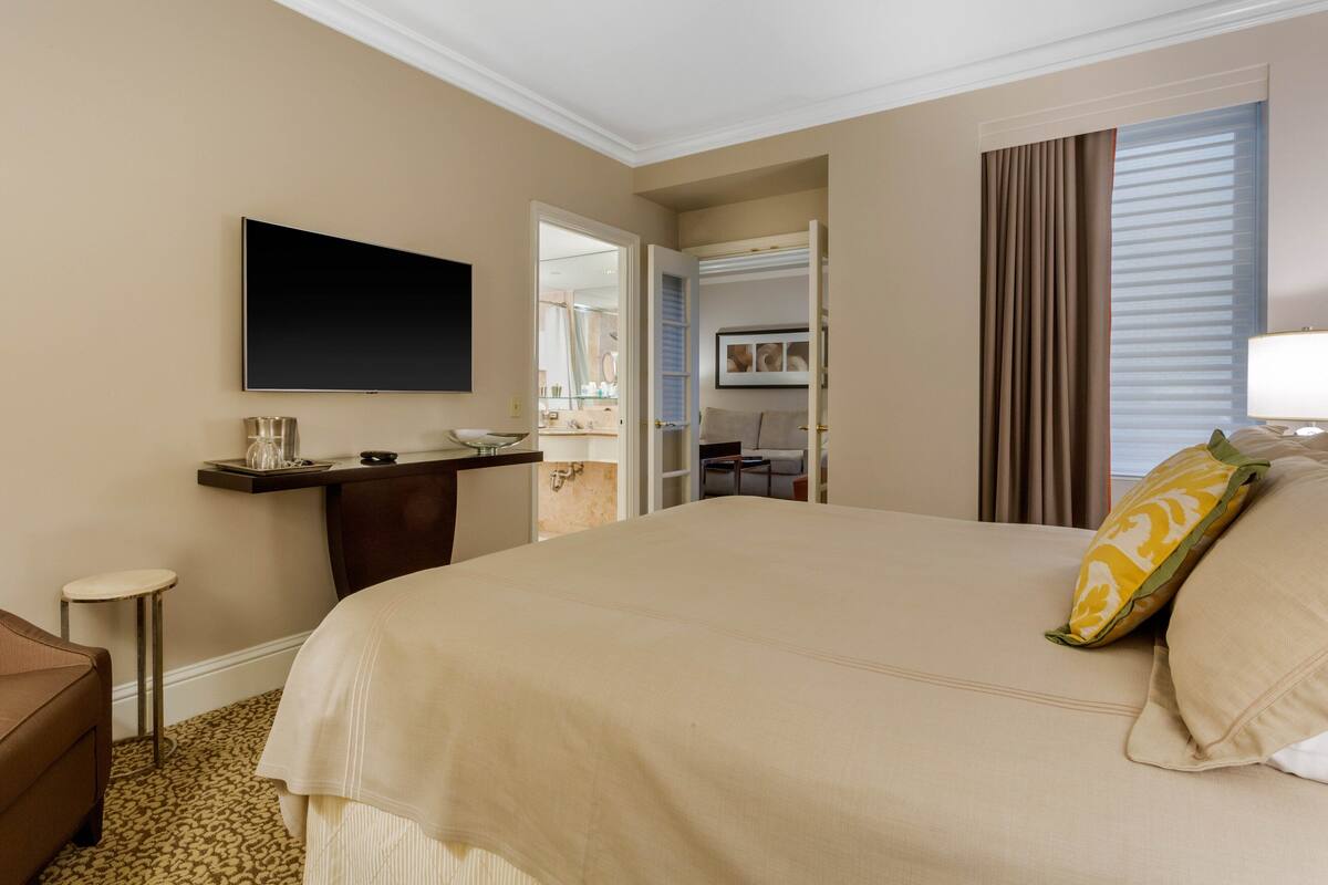 Quarto de hotel com uma cama grande, televisão de tela plana na parede, uma mesinha com luminária e uma cadeira.