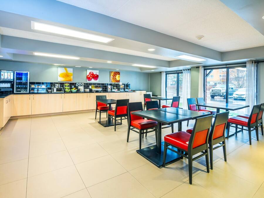 A modern dining area with a long table and red chairs, a counter with food and drink options, and large windows offering a view of the street.