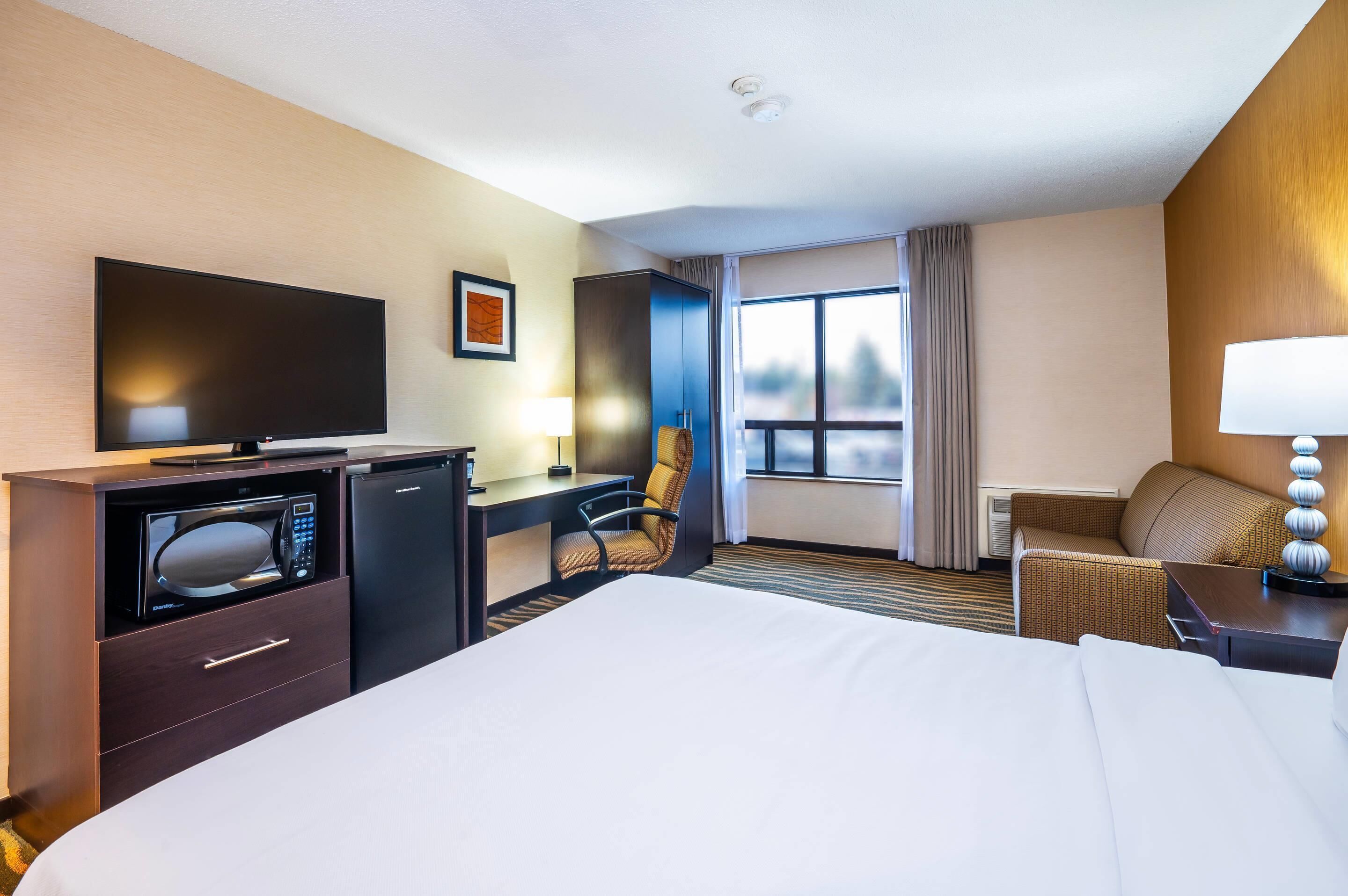 A hotel room with a bed, a desk with a flat-screen TV, a microwave, a chair, a sofa, and a window with curtains.