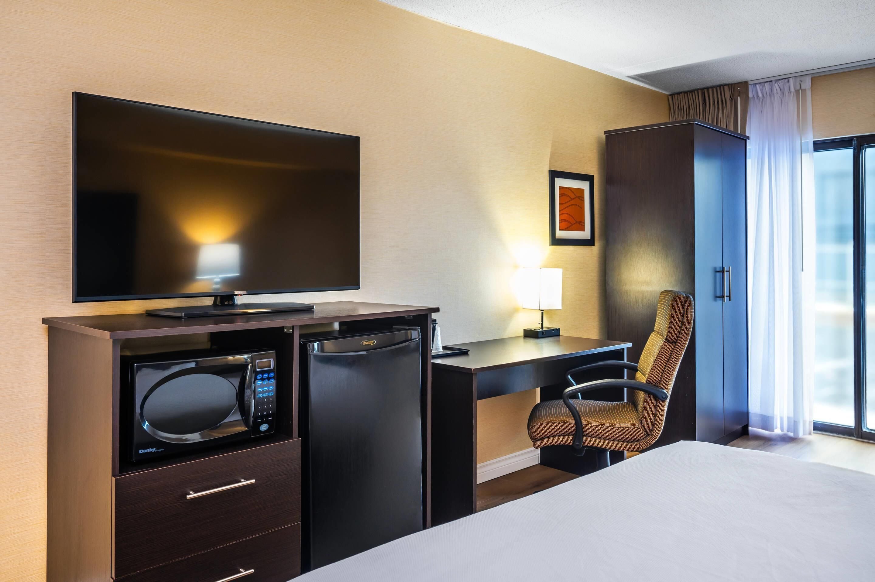 A hotel room with a bed, a desk with a computer, a microwave, a refrigerator, a television, a chair, and a window with curtains.