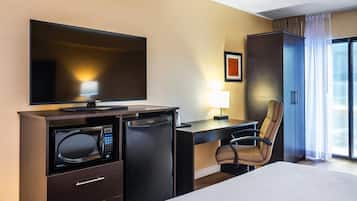 Habitación de hotel con cama, escritorio con computadora, microondas, refrigerador, televisor, una silla y ventana con cortinas.