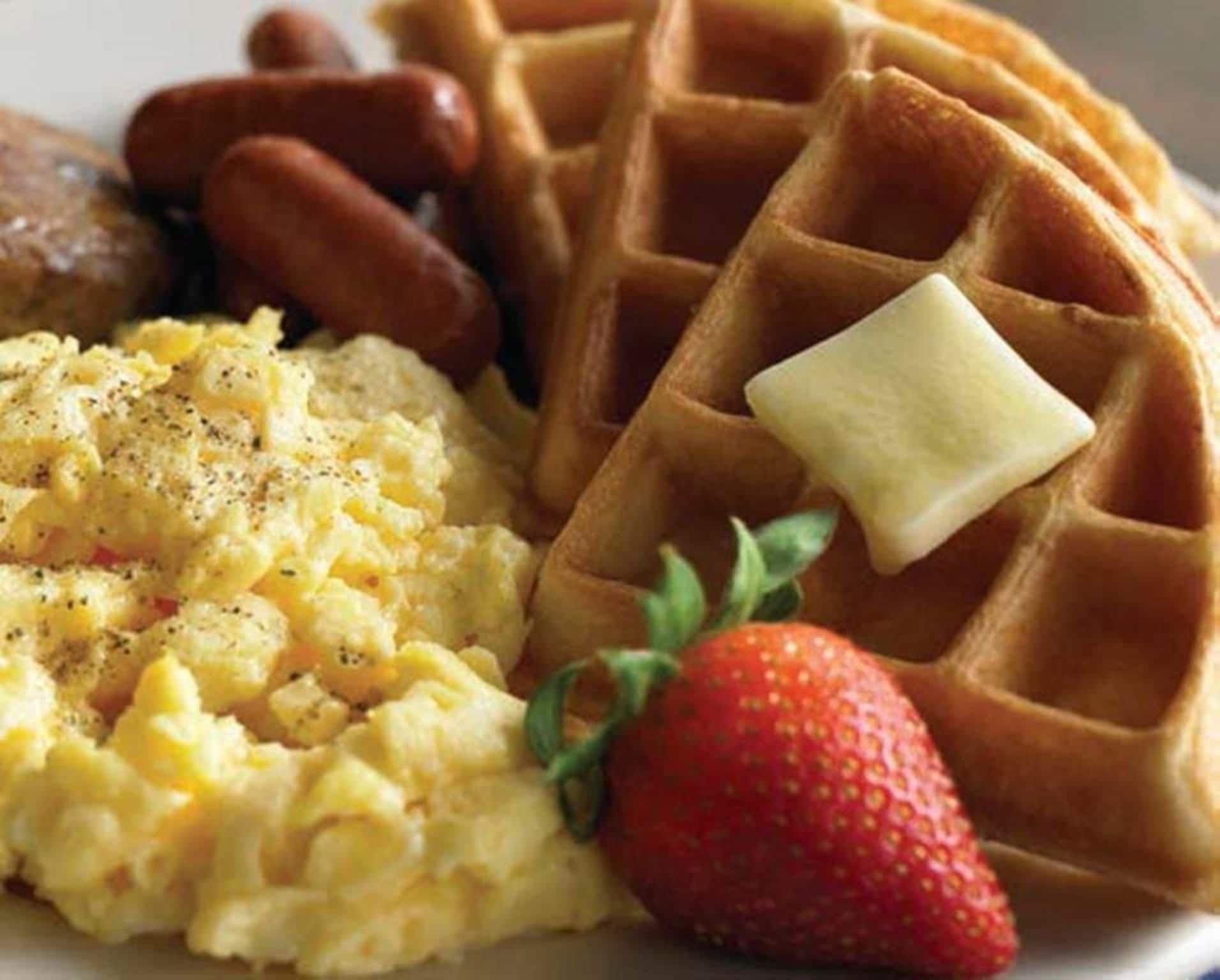 Un plato con huevos revueltos, salchicha, waffles con mantequilla y una fresa.