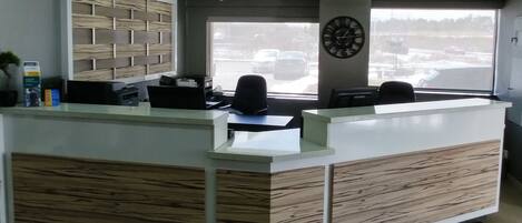 A reception desk with a modern design, featuring a wooden panel front and a glass top. There are two black chairs behind the desk, and a clock is mounted on the wall above.