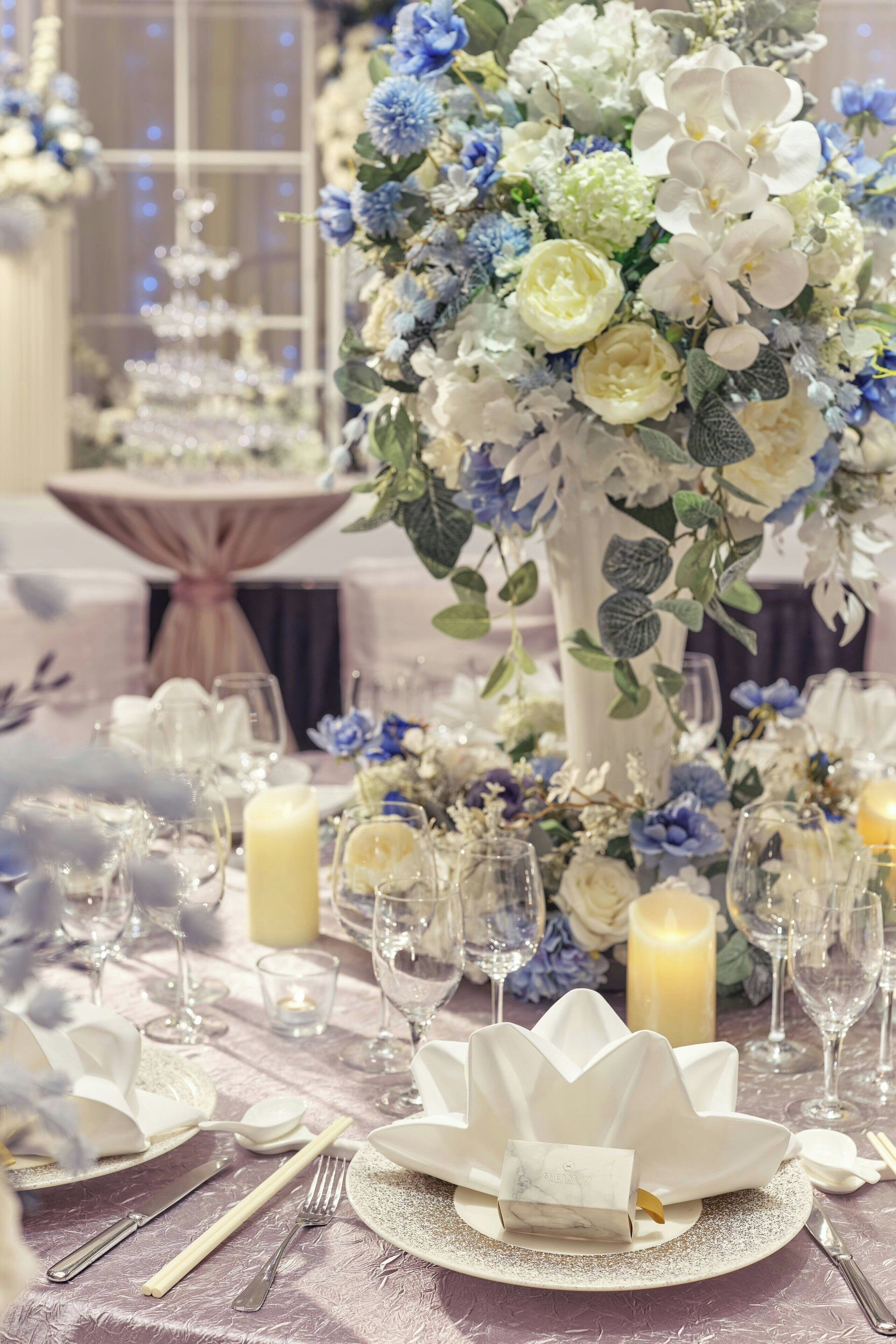 Een gedekte tafel met witte borden, bestek en glazen, omringd door een bloemstuk met witte en blauwe bloemen.
