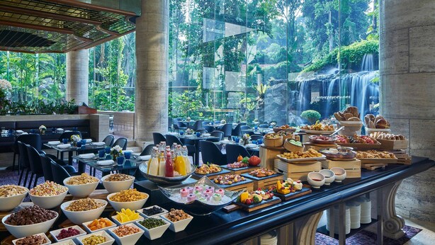 Un bufé con diferentes alimentos y bebidas, ubicado en un salón con amplios ventanales que dan a un jardín exuberante y una cascada.