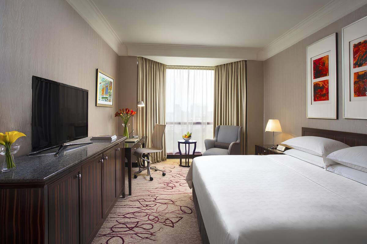 Habitación de hotel con cama, escritorio con televisor, silla, pequeña mesa con lámpara y obras de arte en las paredes.