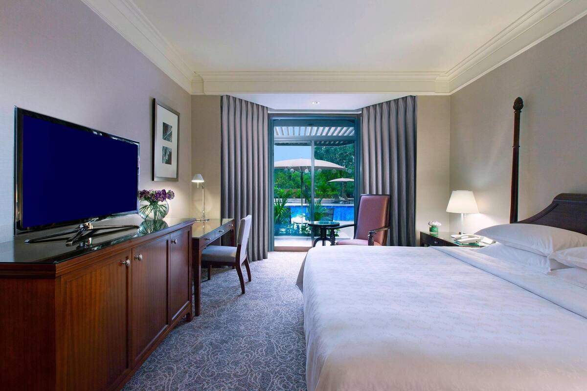 Habitación de hotel con cama, escritorio con televisor, silla, mesita de noche y vista a un espacio exterior con sombrillas.