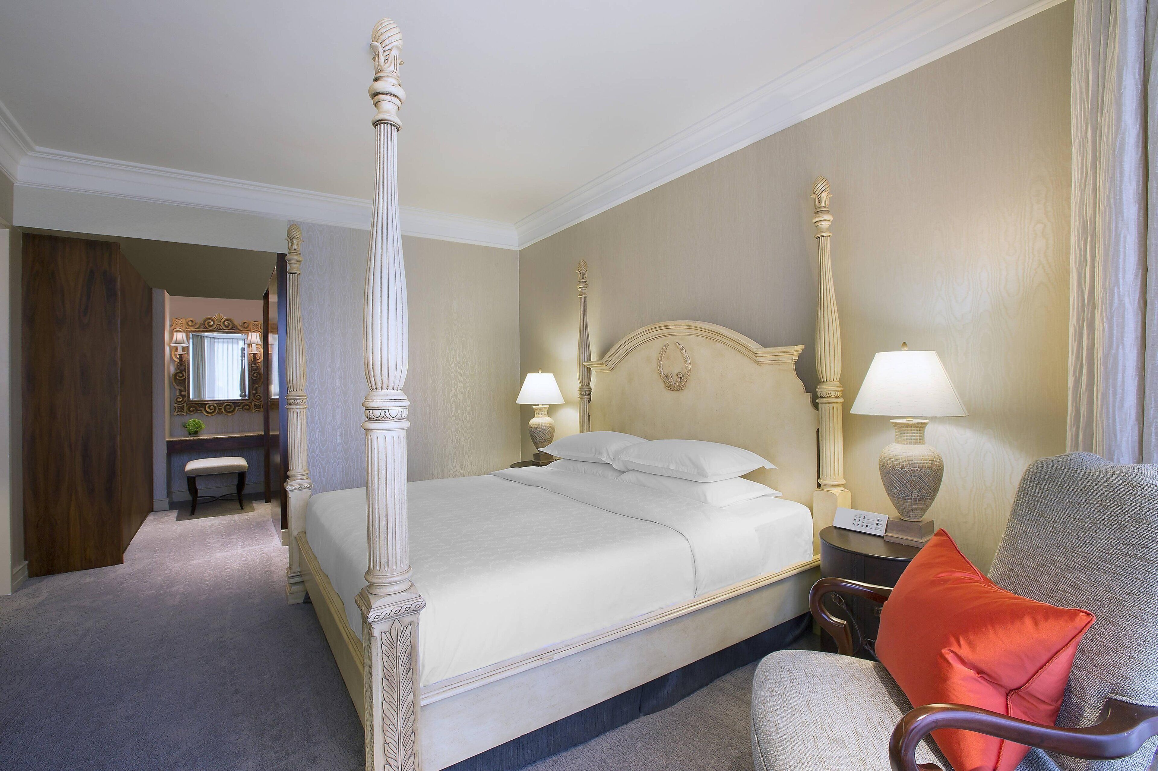 Un lit à baldaquin avec une tête de lit, des lampes de chevet, une chaise avec un coussin rouge et une petite table avec une lampe.
