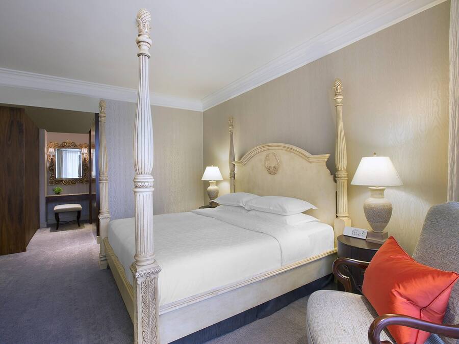 Un lit à baldaquin avec une tête de lit, des lampes de chevet, une chaise avec un coussin rouge et une petite table avec une lampe.