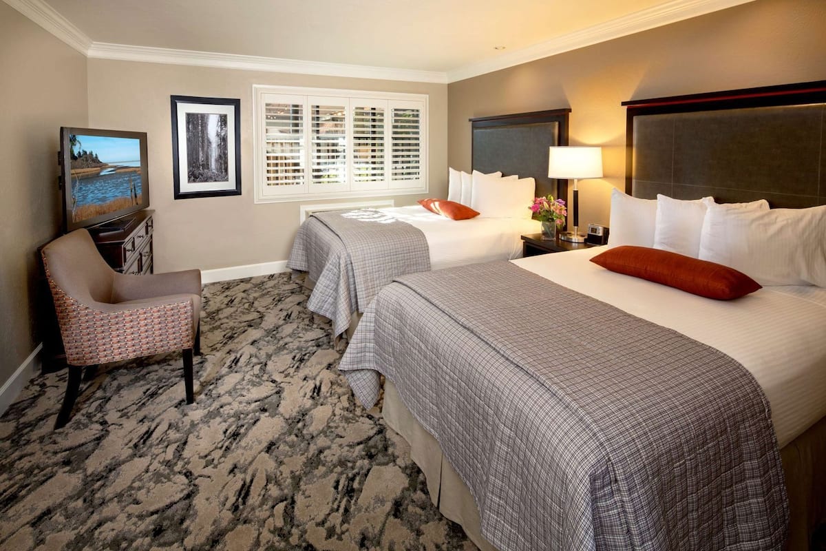 A hotel room with two beds, a chair, a desk, and a window with blinds.