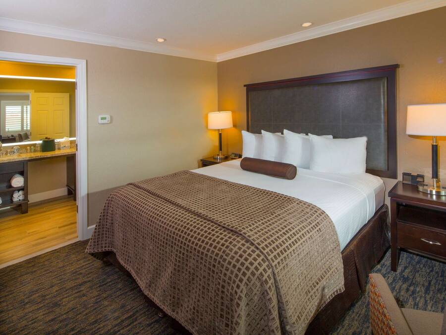 A hotel room with a large bed, two bedside lamps, a nightstand, a chair, and a bathroom visible through an open door.