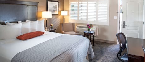 Una habitación de hotel con una cama grande, dos lámparas de noche, una silla, una mesita con un jarrón de flores y una puerta.