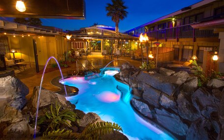 A heated outdoor pool area with a waterfall feature, surrounded by rocks and plants, and adjacent to a building with outdoor seating and a balcony.