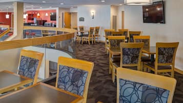 A hotel dining area with multiple tables and chairs, a staircase, and a television mounted on the wall.