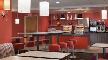 Una cafetería moderna con paredes rojas, diversas zonas de estar que incluyen taburetes y mesas, una máquina de café y una vitrina con pasteles.