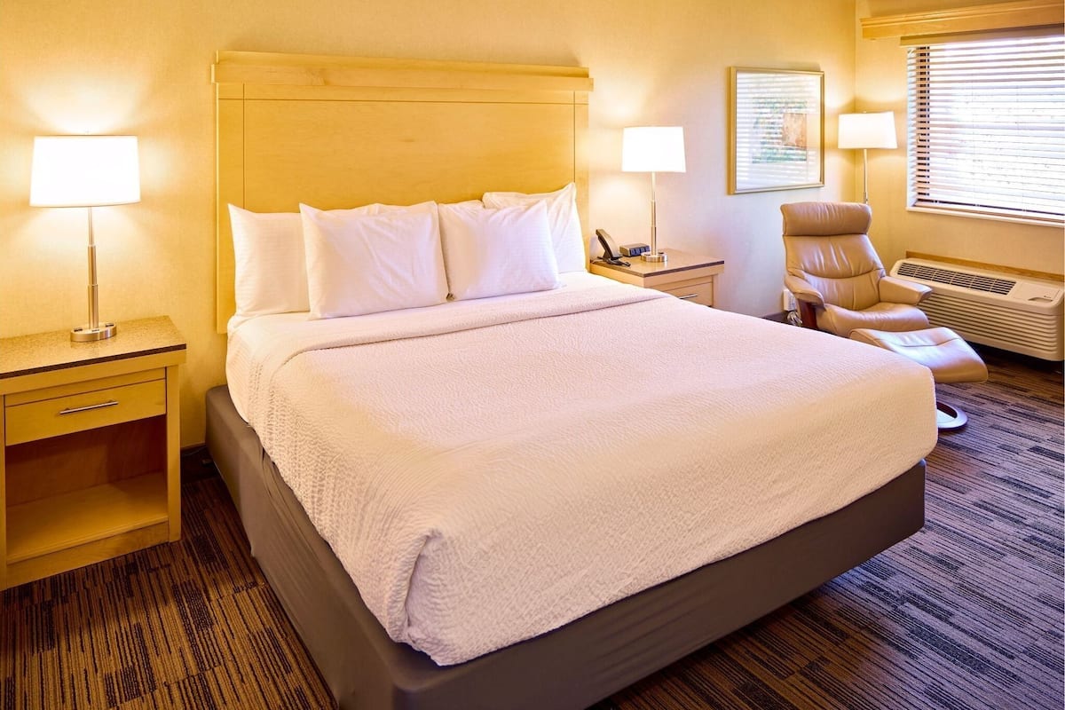Une chambre d’hôtel avec un grand lit, deux tables de chevet, une chaise et une fenêtre avec des stores.