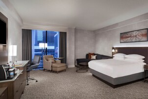 Hotelkamer met een groot bed, een bureau met stoel, een bank en uitzicht op een stadsgezicht door grote ramen.