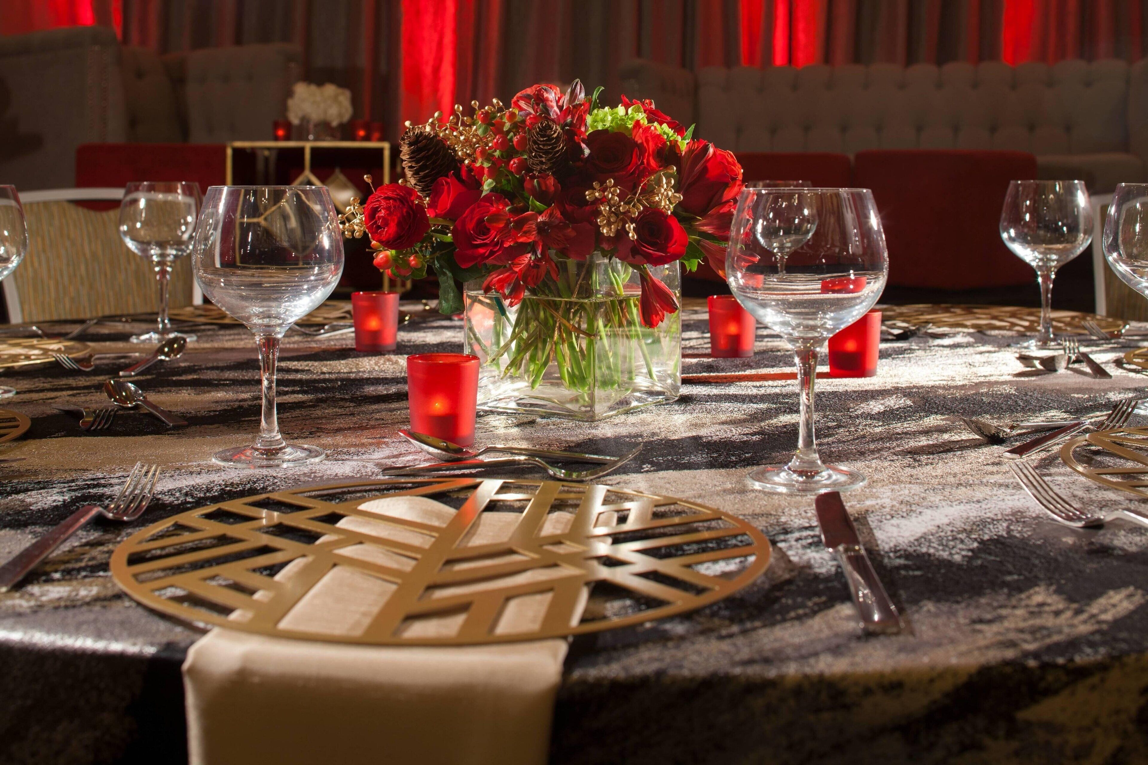 A dining table set with wine glasses, a vase of red flowers, and candles.