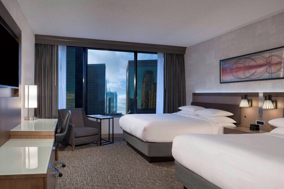 A hotel room with a large bed, a desk, a chair, a TV, and a view of the cityscape through the window.